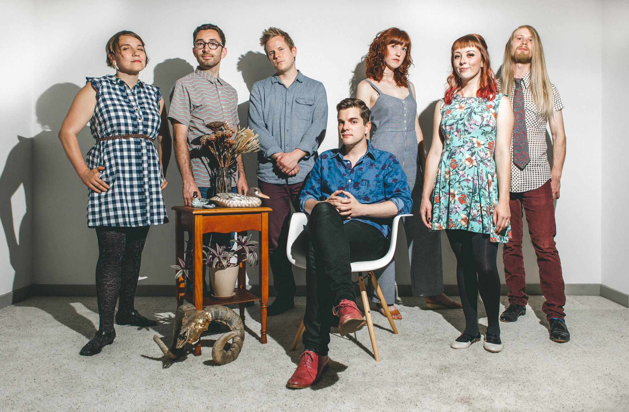 The seven members of Makeunder standing together against a pale wall, with a small wooden side table holding dried flowers, a duck decoy, and a potted plant, and a ram skull on the floor.